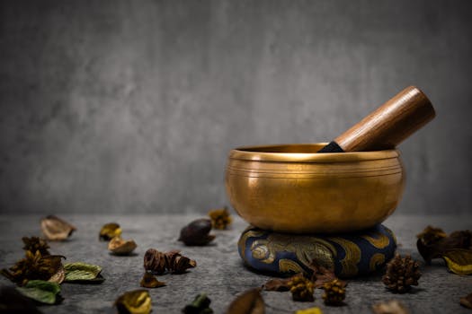 Golden tibetan singing bowl standing on a small blue cushion with wooden stick and dried plants scattered around, promoting relaxation and meditation against a gray backdrop